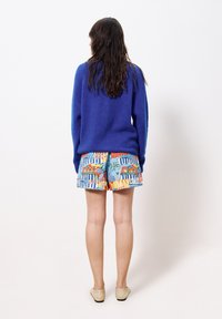 Femme aux longs cheveux bruns portant un pull bleu, un short à motifs colorés et des ballerines beiges, se tenant face à un mur blanc.