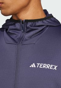 Haut de sweat à capuche zippé bleu marine avec un col haut, fabriqué en tissu léger. Présente une bordure noire et un logo "TERREX" blanc sur la poitrine.