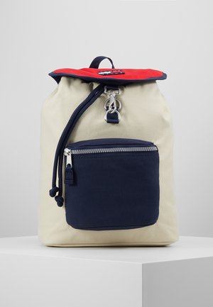 Beige rugzak met een rood klepje, marineblauwe voorzak met rits, trekkoordsluiting en metalen gesp, geplaatst op een witte sokkel tegen een grijze achtergrond.