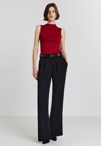 Top rosso senza maniche con un collo alto, abbinato a pantaloni neri a gamba larga con vita alta, dotati di una cintura con decorazioni in argento.