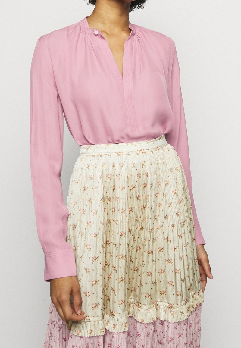 Rosa långärmad blus med en krage med knäppning, i kombination med en plisserad krämfärgad kjol med små blommönster och volangkant.