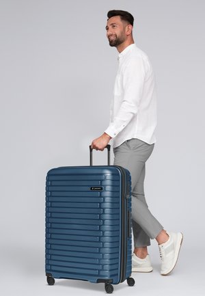 Valigia rigida blu con texture a righe verticali, doppie ruote e manico retrattile, mostrata accanto a un uomo in abbigliamento casual.