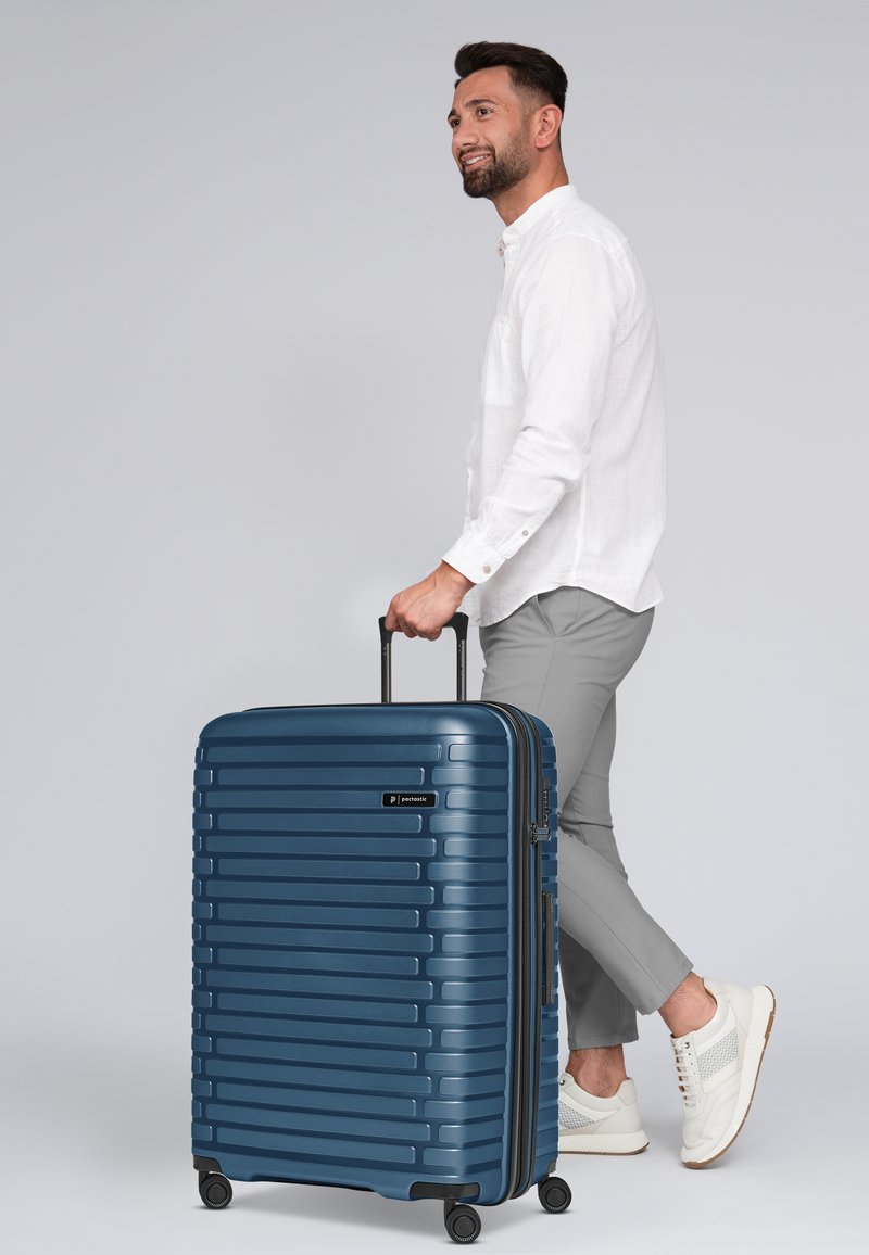 Maleta rígida azul con textura de surcos verticales, ruedas dobles y asa retráctil, mostrada junto a un hombre en atuendo casual.