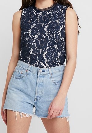 Granatowy top bez rękawów z kwiatowej koronki w zestawieniu z jasnoniebieskimi, wysokim stanem, dżinsowymi szortami z postrzępionymi brzegami i zapięciem na guziki z przodu.