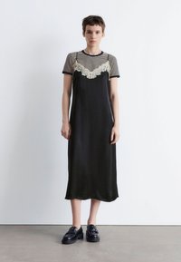 Schwarzes Satin-Kleid mit Spitze, über einem kurzärmeligen, navy-weißen gestreiften Hemd geschichtet. Das Model trägt schwarze Plateauschuhe.