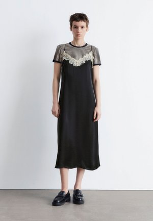Schwarzes Satin-Kleid mit Spitze, über einem kurzärmeligen, navy-weißen gestreiften Hemd geschichtet. Das Model trägt schwarze Plateauschuhe.
