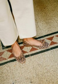 Des ballerines imprimées léopard avec un bout rond, un nœud sur le devant et une surface texturée, associées à un pantalon à jambes larges de couleur crème sur un sol à motifs.