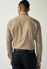 Vaalean beige pitkähihaisessa paidassa on muotoiltu kaulus ja napitetut hihansuut, yhdistettynä tyylikkäisiin tummiin housuihin ja mustaan vyöhön.
