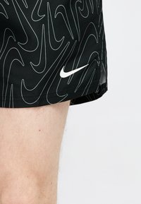 Μαύρο αθλητικό σορτς με λευκά αφηρημένα γραμμικά σχέδια και λευκό λογότυπο Nike swoosh, φορεμένο σε ελαφρώς τριχωτό πόδι.