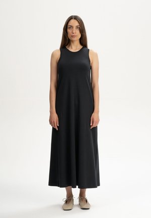 Femme aux longs cheveux bruns portant une robe maxi noire sans manches et des chaussures plates beiges, debout devant un fond blanc uni.