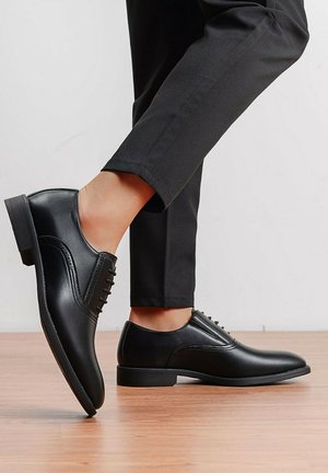 Chaussures habillées en cuir noir portées avec un pantalon noir, un pied levé, l'autre posé à plat sur un sol en bois, sur un fond clair uni.