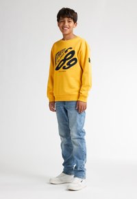 Sudadera amarilla con estampado gráfico negro, jeans azules y zapatillas blancas. La sudadera tiene cuello redondo y puños acanalados.
