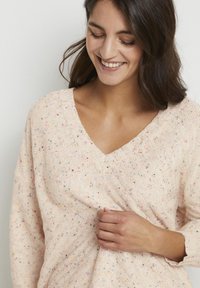 Maglione lavorato a maglia rosa chiaro con scollo a V, maniche lunghe e macchie multicolori su tutto il tessuto, che evidenziano un design testurizzato.