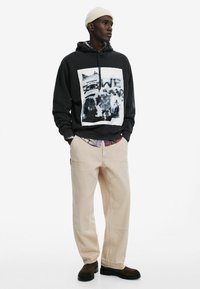 Felpa nera con stampa grafica, pantaloni bej larghi e scarpe scure. Il modello indossa anche un berretto crema e una camicia aggiuntiva sotto.