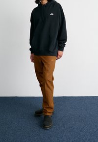 Černá mikina s kapucí s předním kapsou a bílým logem, doplněná o hnědé kalhoty s odpovídajícím logem. Černá obuv s texturovanou podrá�žkou.