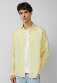 Chemise boutonnée jaune clair en tissu doux, avec des manches longues, un col et une coupe décontractée, associée à un T-shirt blanc.
