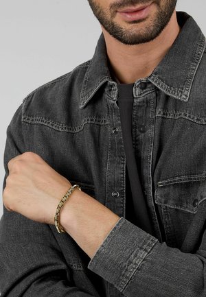 Bracciale in argento e oro composto da catene metalliche intrecciate, indossato al polso, con una giacca di jeans testurizzata in grigio scuro e una maglietta nera sotto.