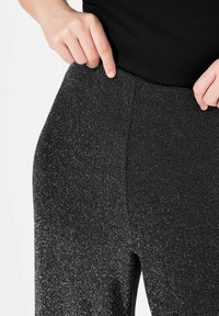Pantalons en texture métallique noire avec un subtil éclat. Design ajusté avec une taille élastique. Gros plan montrant le détail du tissu et les coutures.