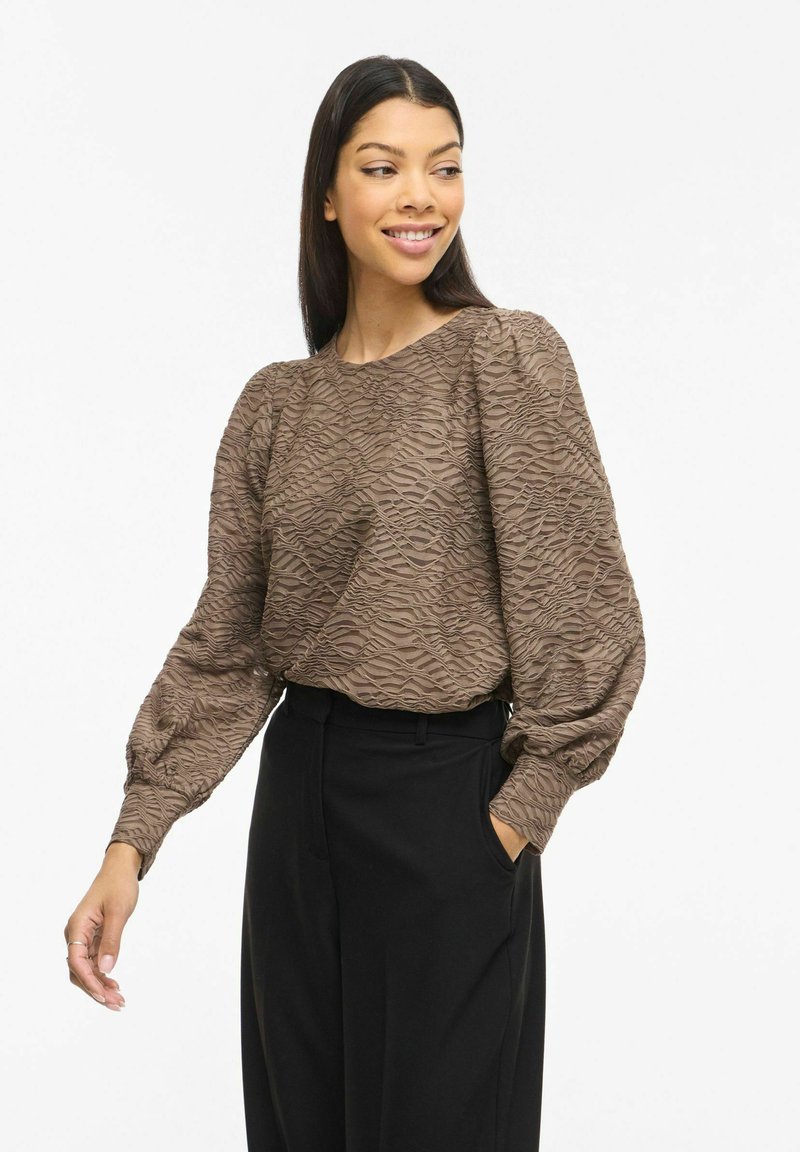 Blouse marron texturée avec un motif ondulé, manches longues bouffantes et col rond, associée à un pantalon noir taille haute.