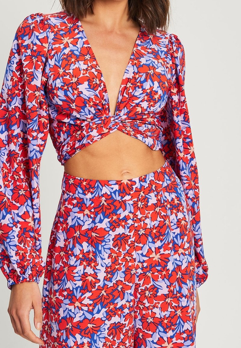 Femme portant un crop top à manches longues fleurie rouge, violet et rose avec un devant torsadé et un pantalon taille haute assorti.