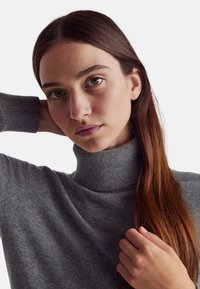 Šedý pletený svetřík s rolákem, který se vyznačuje měkkou texturou a volným střihem, s žebrovaným výstřihem a detaily na ramenou. Vlasy rámují obličej.