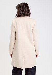 Manteau en laine beige à manches longues et coupe droite, avec un détail de couture au dos. Tissu texturé, conception longueur genoux. Sans fermetures visibles.