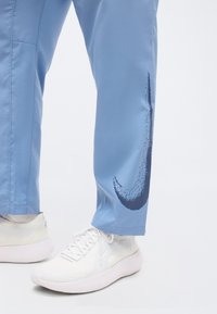 Κάτω μέρη ποδιών που φορούν ανοιχτό μπλε παντελόνια με ένα μεγάλο σκοτεινό σχεδιασμό σβούρας κοντά στον αστράγαλο και λευκά αθλητικά παπούτσια με κορδόνια σε λευκό φόντο.