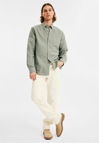 Groene button-up shirt met een voorzak, gedragen met crèmekleurige broek en beige schoeisel. Natuurlijke stoftextuur is zichtbaar.