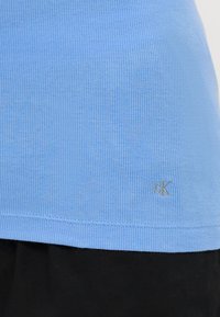 Light blue ribbed T-shirt, short sleeves, featuring a small embroidered logo at the hem. Fabric texture is smooth and stretchy.