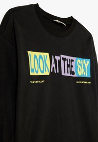 Camiseta negra con texto audaz y colorido: "MIRA AL CIELO" en azul, amarillo, púrpura y verde. Tejido suave con un corte relajado.