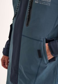 Un gros plan de la main d'une personne dans une veste de sport bleu marine, mettant en valeur une poche zippée et des détails en tissu texturé.