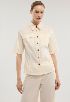 Frau trägt ein kurzärmeliges cremefarbenes Hemd mit zwei Brusttaschen und passende cremefarbene Hosen vor einem einfarbigen Hintergrund.