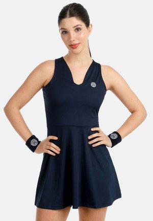 Mujer con cabello oscuro recogido, vestida con un vestido azul marino sin mangas y pulseras a juego, de pie con las manos en las caderas contra un fondo liso.
