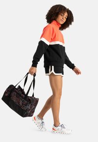 Color-blocked fleece hoodie in black, orange, and white, paired with black shorts and a black duffel bag with colorful patterns. White sneakers.