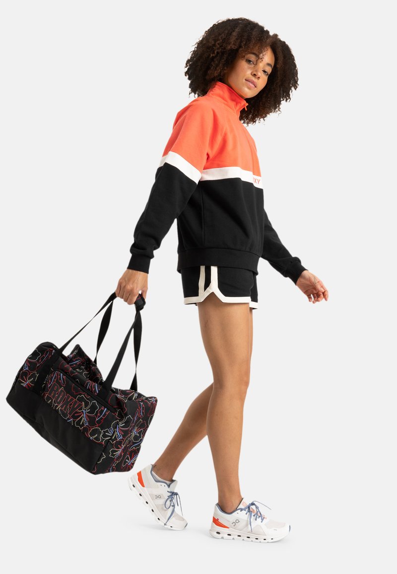 Color-blocked fleece hoodie in black, orange, and white, paired with black shorts and a black duffel bag with colorful patterns. White sneakers.