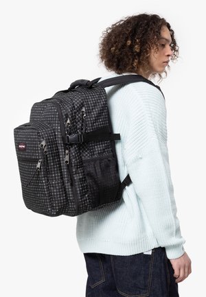 Mochila a cuadros en blanco y negro con múltiples bolsillos con cremallera, correas ajustables, tela texturizada y bolsillo lateral de malla.