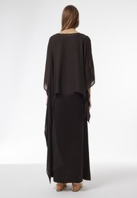 Langes schwarzes Kleid mit einem fließenden, transparenten Überwurf. Verfügt über einen runden Ausschnitt und weite Ärmel, kombiniert mit flachen Sandalen.