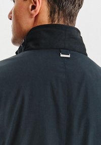 Granatowa kurtka z wysokim kołnierzem, gładką fakturą i małą metką z logo widoczną przy szwie kołnierza. Prosty, funkcjonalny design.