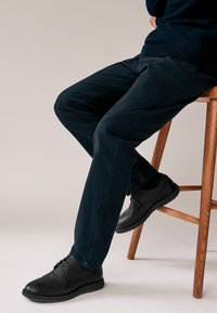 Pantalon bleu marine à texture lisse, associé à des chaussures en cuir noir avec lacets et semelle en caoutchouc, reposant sur un tabouret en bois.