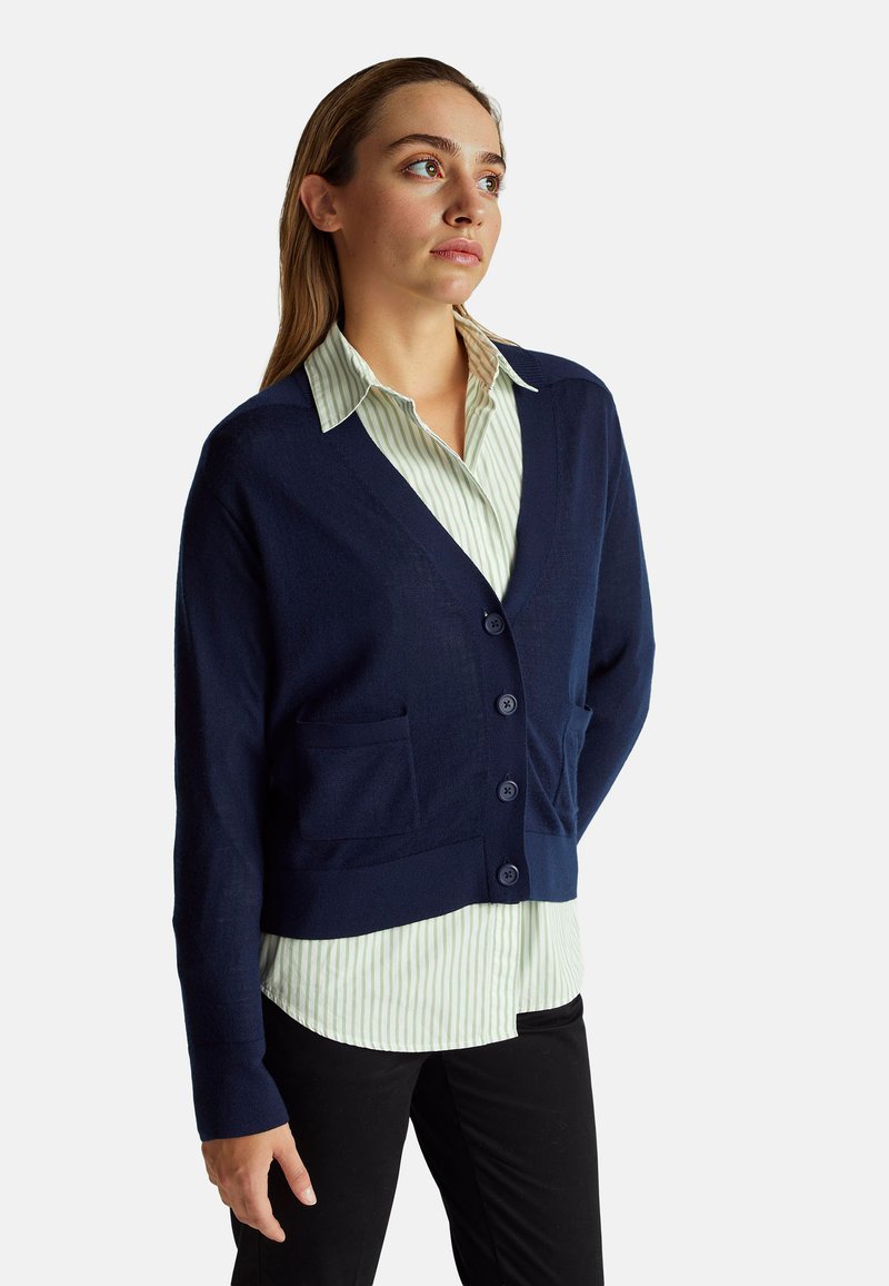 Granatowy kardigan z dekoltem w serek, z pięcioma guzikami i dwoma przednimi kieszeniami, noszony na jasnoszarej koszuli w paski. Miękka struktura dzianiny.