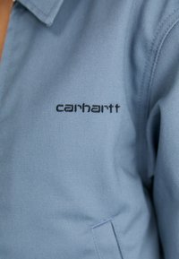 Giacca azzurro chiaro realizzata in un tessuto resistente. Presenta un colletto a punta e un logo "Carhartt" ricamato in nero sul davanti. Design semplice e funzionale.