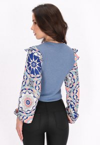 Maglione a maniche lunghe blu a coste con maniche floreali a motivi in rosa e blu marin. Presenta dettagli arricciati sulle spalle. Pantaloni neri aderenti.