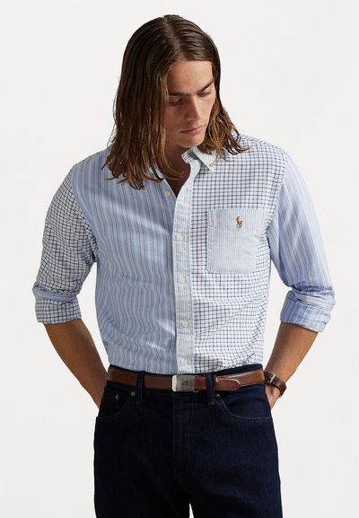 Homme aux longs cheveux portant une chemise boutonnée à carreaux et rayures bleues et blanches en patchwork, un jean noir et une ceinture en cuir marron avec une boucle en argent.