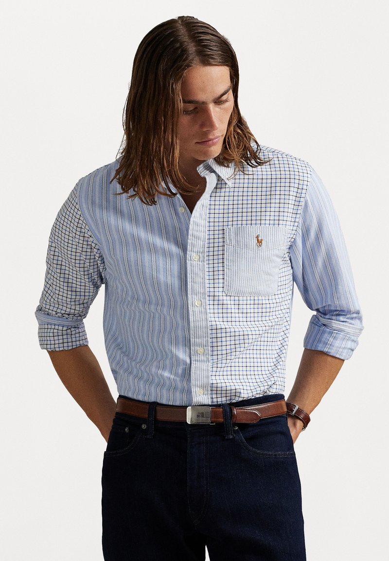 Man met lang haar in een patchwork blauw-wit geruite en gestreepte overhemd met knopen, donkere spijkerbroek en een bruine leren riem met een zilveren gesp.
