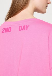 Camiseta de algodón rosa con un gran y audaz texto "2ND DAY" impreso en la parte de atrás. La tela tiene una textura suave y un diseño de corte relajado.