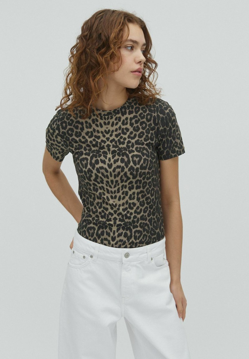 Jeune femme aux cheveux bouclés portant un t-shirt à motif léopard et un pantalon blanc, regardant sur le côté sur un fond uni.