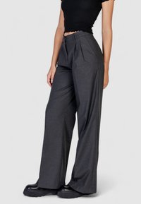 Pantaloni grigio carbone plissettati con una vestibilità sartoriale, realizzati in un tessuto testurizzato. Presentano una chiusura con zip e un design a gamba larga. Abbinati a scarpe con piattaforma nere.