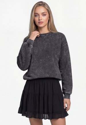 Młoda kobieta o długich blond włosach, ubrana w wyprany czarny, oversize’owy sweter oraz czarną warstwową mini spódniczkę na białym tle.
