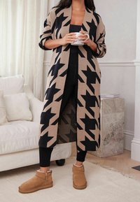 Nainen seisoo sisätiloissa beigeä ja mustaa kuvioitua pitkää neuletakkia, mustia legginssejä ja vaaleanruskeita talvikenkiä käyttäen, pitäen valkoista mukia.