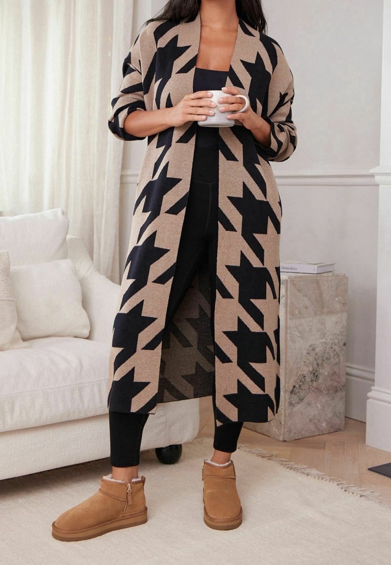 Nainen seisoo sisätiloissa beigeä ja mustaa kuvioitua pitkää neuletakkia, mustia legginssejä ja vaaleanruskeita talvikenkiä käyttäen, pitäen valkoista mukia.